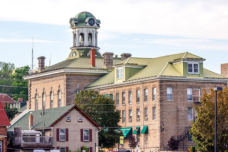  A stone building with hip roofs, dormers, and a tall clock tower amongst trees and neighbouring houses.